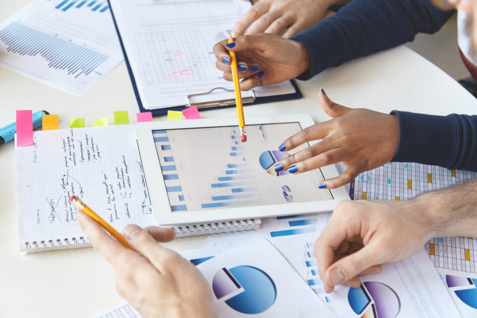 Colleagues working together on financial report using modern gadget. African girl with pencil presenting information on laptop screen, studying company's growth, showing diagram to her partners