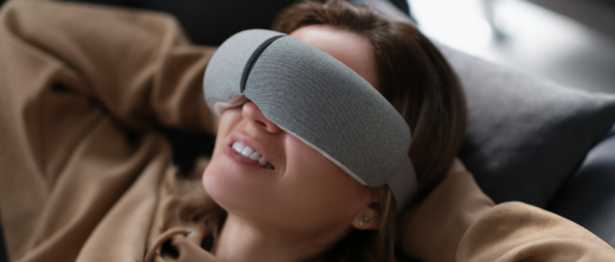 A woman relaxing on a couch with a gray eye mask and using an eye massager.
